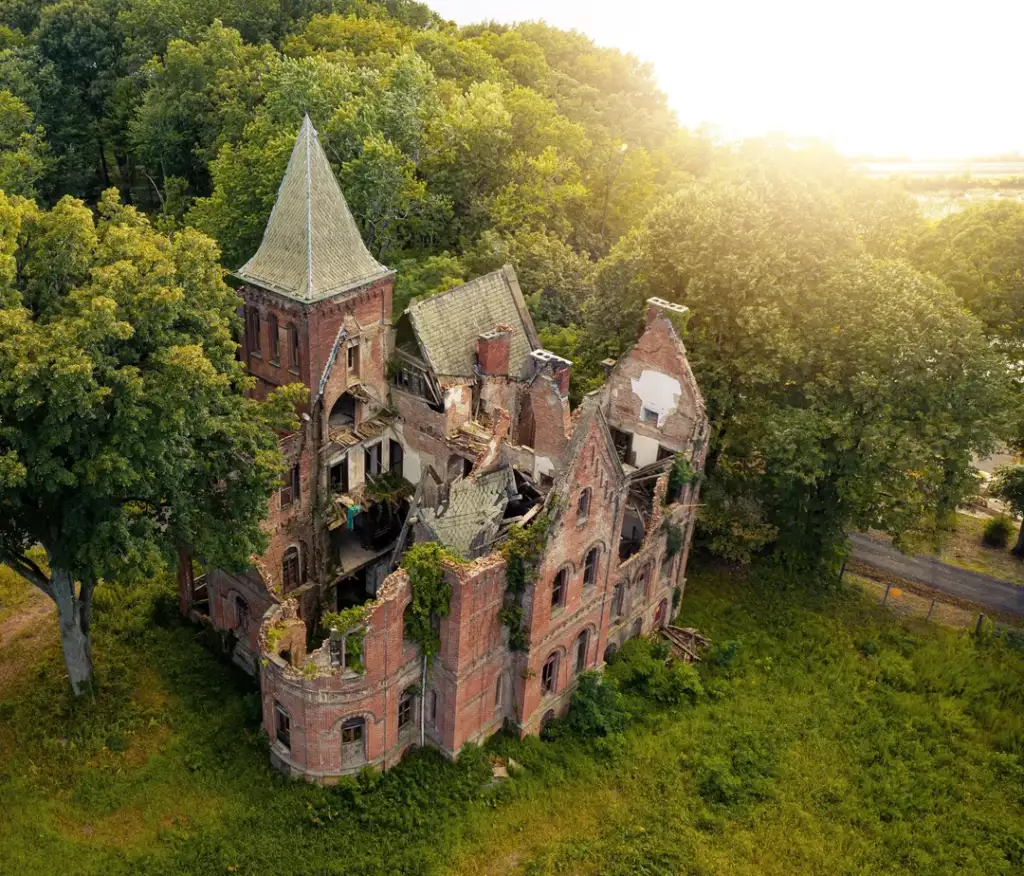 An aerial view of a large, abandoned brick building with a collapsed roof and overgrown vegetation. The building has a prominent pointed tower and is surrounded by lush green trees. Sunlight filters through the trees, illuminating the scene.