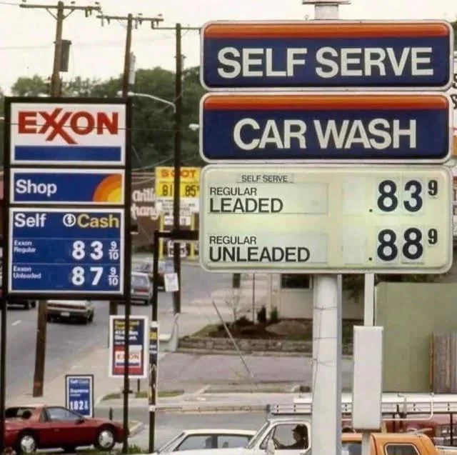 A vintage photo of an Exxon gas station with a sign showing fuel prices: self-serve regular unleaded at 83.9 cents and regular unleaded at 88.9 cents. Adjacent, another sign advertises a car wash. Power lines and road signs are visible in the background.