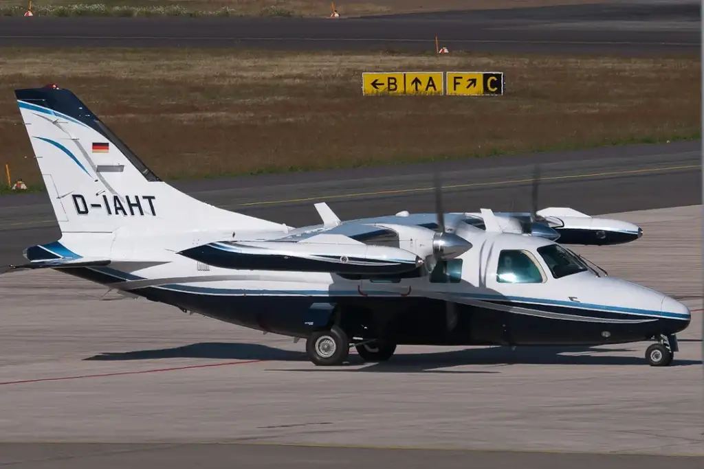 A small twin-engine aircraft with a white and blue exterior is parked on an airport runway. The tail number "D-IAHT" is visible. The background shows grassy areas and airport signage.