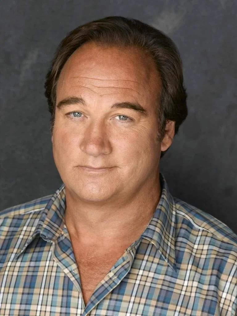 Man with short, dark hair wearing a plaid shirt against a plain, dark background, looking at the camera with a slight smile.