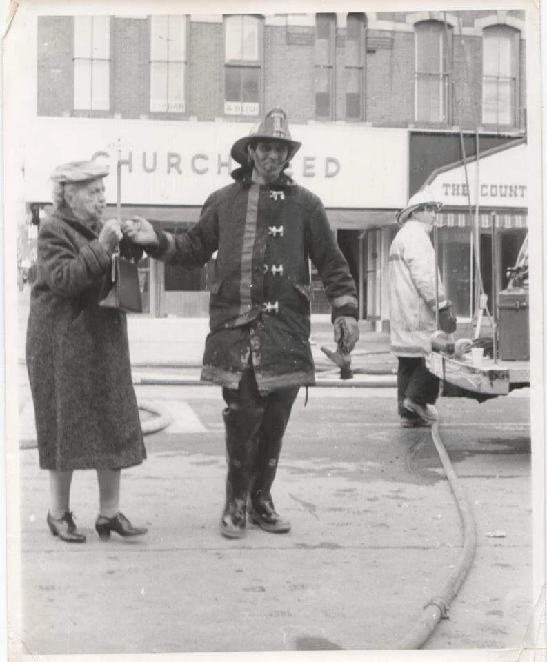 A firefighter in uniform helps an elderly woman on a street, with hoses on the ground. Another firefighter stands near equipment in the background. Buildings with visible signs are in the background. The scene appears to be from a bygone era.