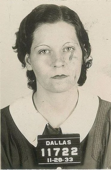 Black and white mugshot of a woman with short, dark hair and a neutral expression. She wears a dark top with a white collar. Her plaque reads "DALLAS 11722 11-28-33," and she has a visible mark on her face.