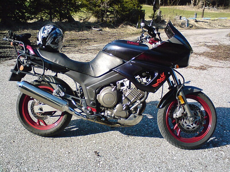 A black Yamaha motorcycle with red rims is parked on a gravel surface. It features a large front fairing and a helmet resting on the seat. The backdrop shows some grass and trees.