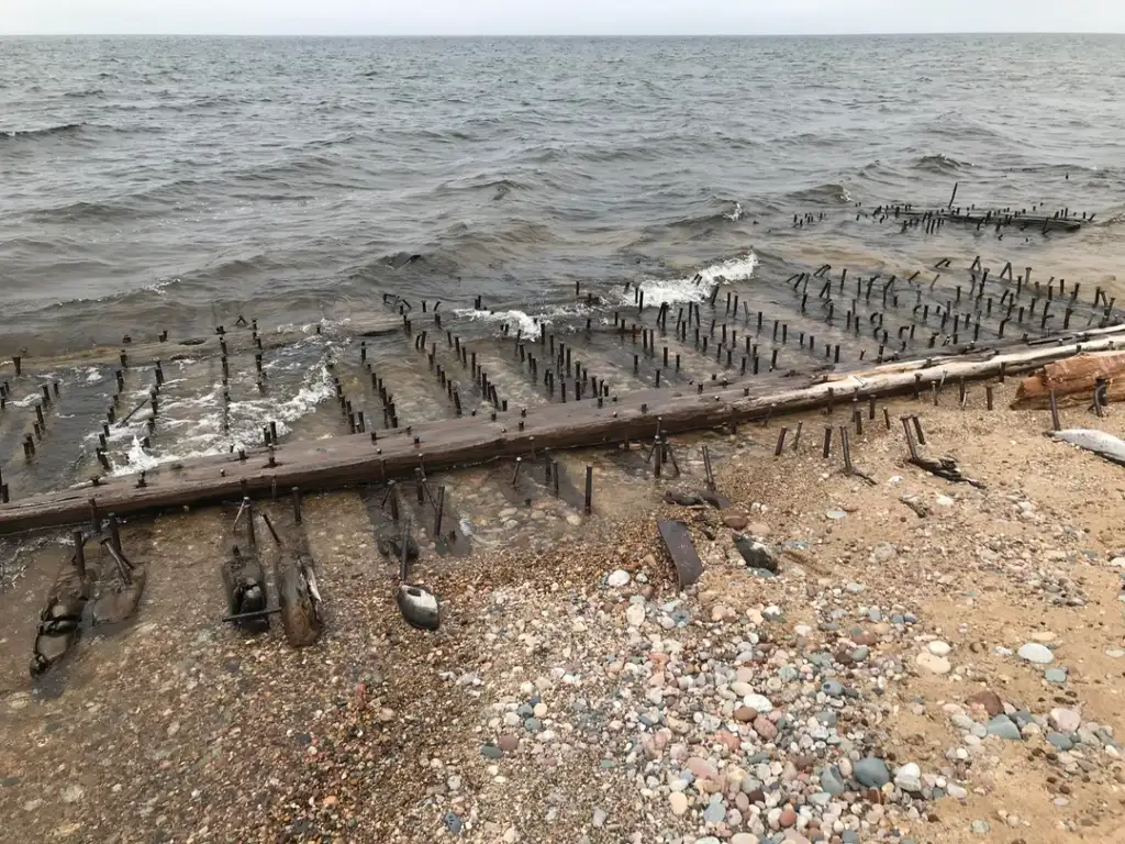 Waves wash over a weathered wooden structure with rusty spikes protruding, partially submerged in a pebbly beach along a vast body of water under an overcast sky.