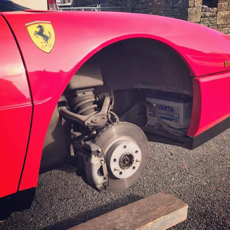 Close-up of a red sports car with the front left wheel removed, revealing the suspension and brake disc. A Varta battery is visible in the wheel well. The car's logo is displayed on the fender. The vehicle is supported by a piece of wood.