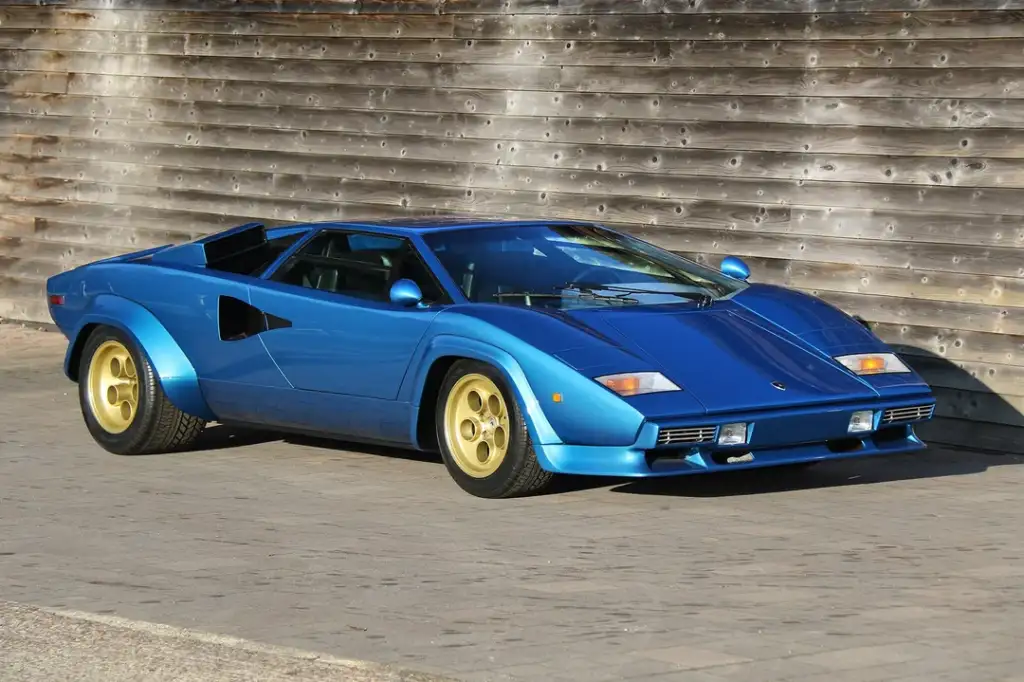 A blue sports car with angular design and gold rims is parked on a paved surface. The background features a weathered wooden wall, enhancing the car's sleek, retro aesthetic.