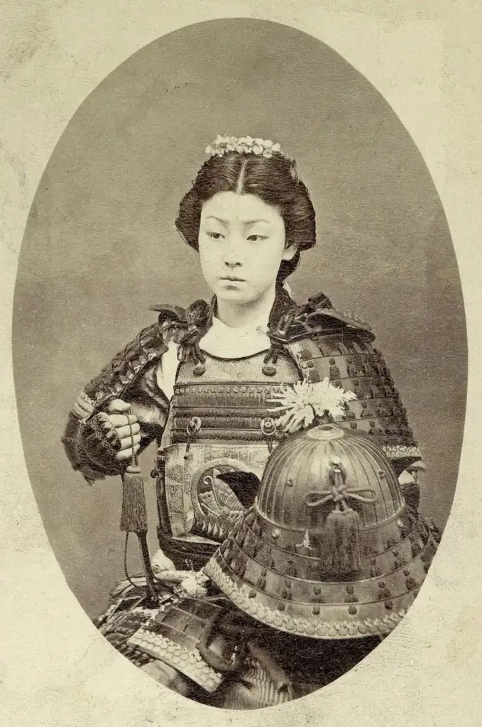 A vintage sepia-toned photograph of a young woman in traditional samurai armor, holding a helmet. She has an ornate hairstyle and wears a determined expression, set against a plain, oval backdrop.