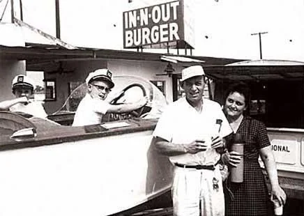 A vintage photo of three men in a drive-thru at In-N-Out Burger. Two men are in a car, one in uniform, while a man in a cap and woman stand beside the car holding drinks. The In-N-Out Burger sign is visible in the background.