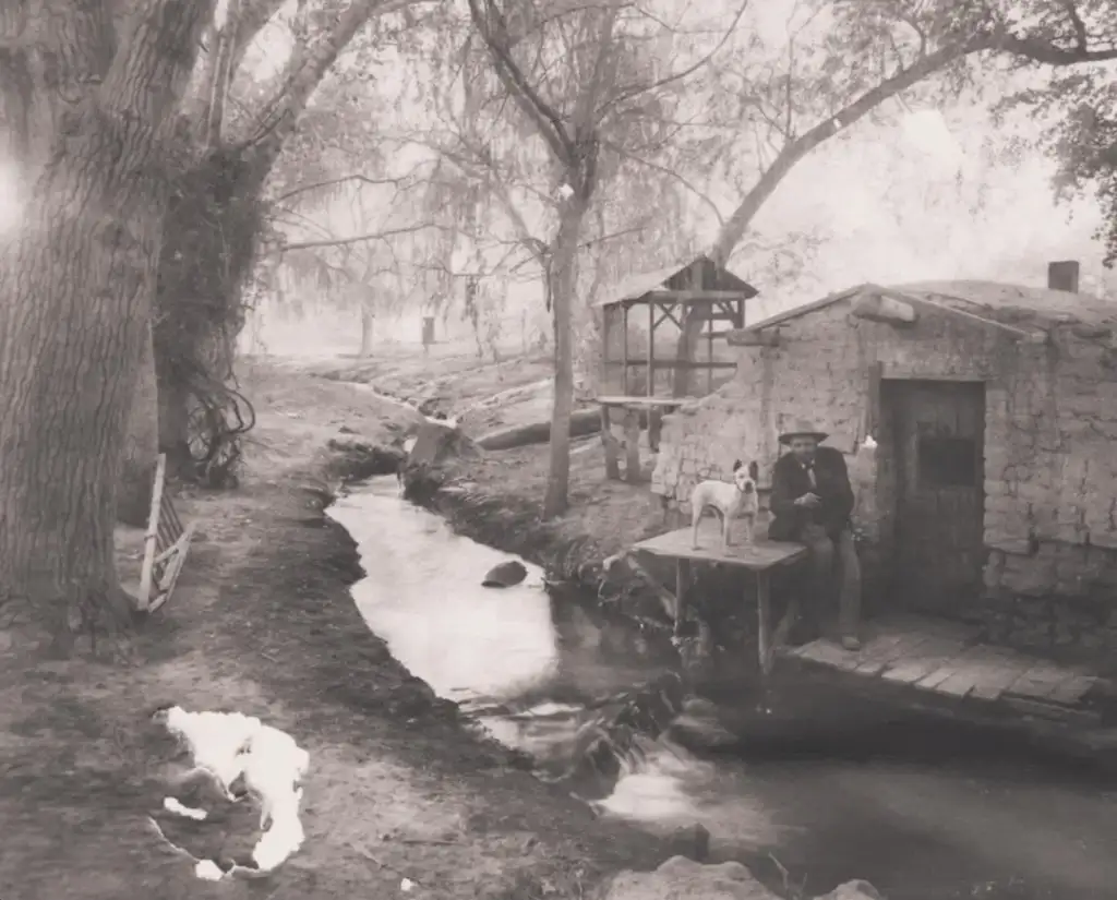 A man sits by a small, rustic stone building near a stream, holding a dog on a wooden platform. The scene is set in a forested area with large, bare trees and a wooden structure in the background, captured in black and white.