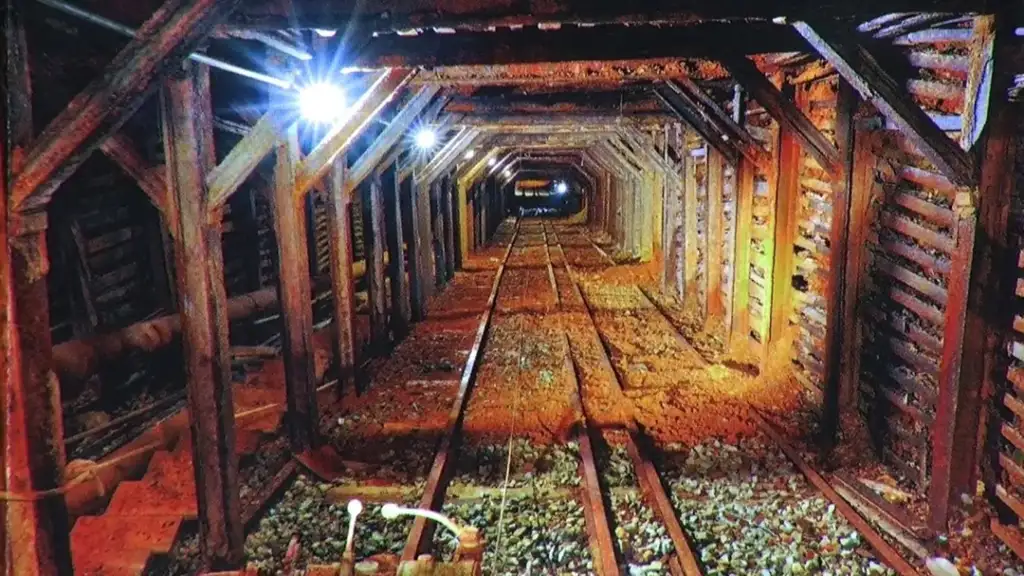 A dimly lit underground mine shaft with wooden supports and reinforced walls. The tracks run along the center, leading into the distance. Bright lights illuminate sections of the tunnel, highlighting the aged and rusty surroundings.