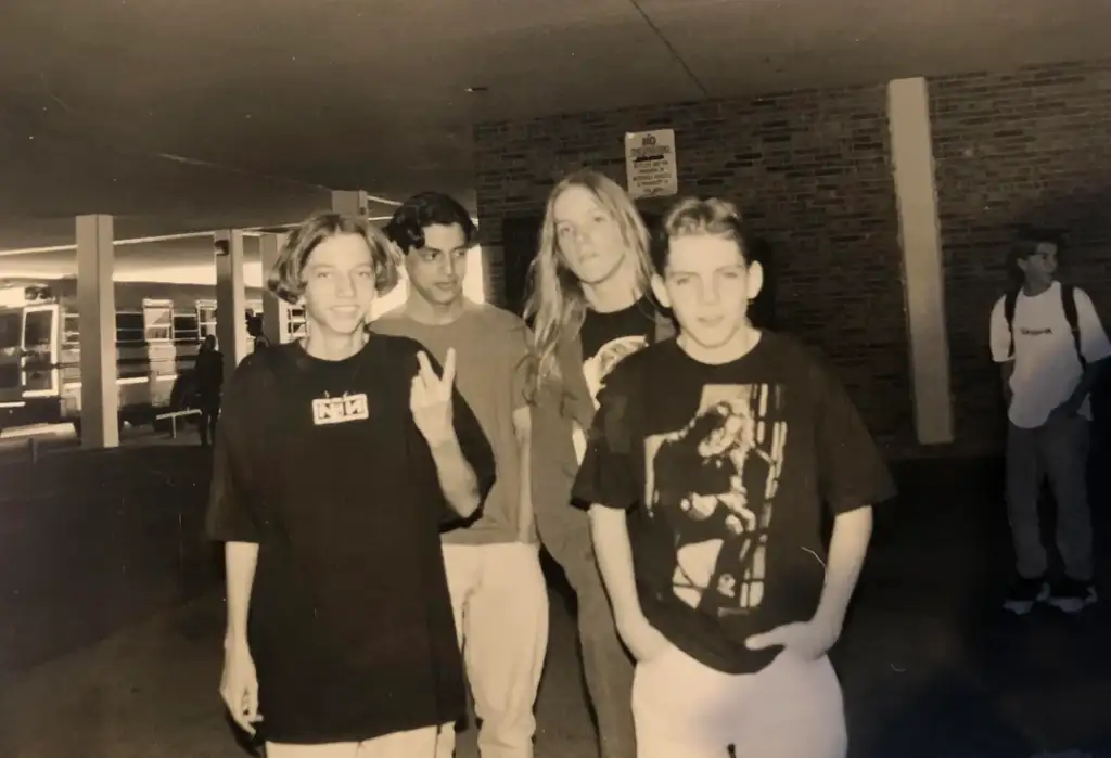 Four young people walk together in a black-and-white photo. One person flashes a peace sign. They all wear casual clothing, and the setting appears to be outdoors near a building. Another person stands in the background.