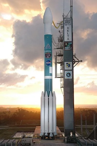 A Delta II rocket stands vertically on a launch pad at sunset, with various logos, including NASA's, visible on its body. The sky is partly cloudy, casting dramatic light on the scene.