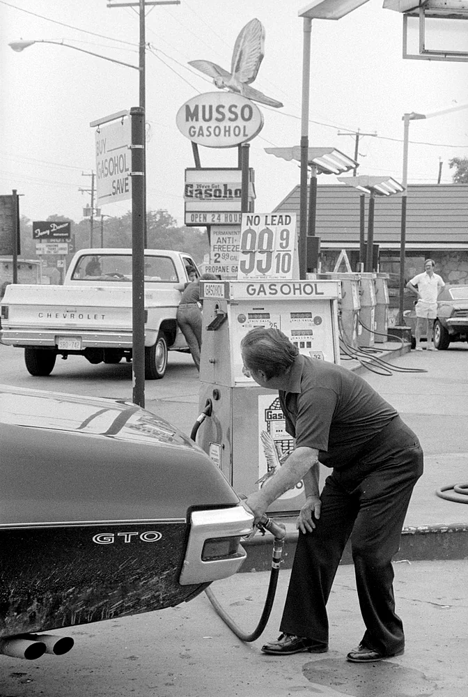 A man fills a classic GTO car with gasohol at a vintage Musso gas station. A Chevrolet truck and other vehicles are visible. Signs display gas prices and promotions. Another person is in the background near the pumps.