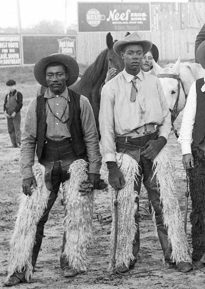 Two cowboys wearing hats and chaps stand in front of horses in a black-and-white photo. They appear focused, with their hands resting by their sides. In the background, a horse and additional figures are visible near a building with signage.