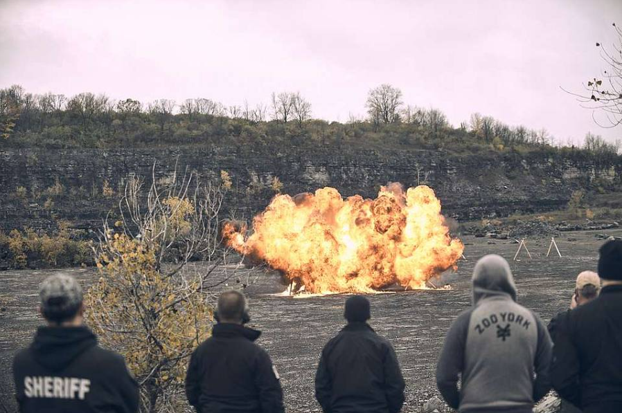 Several people in jackets watch a large explosion in an open, barren area. The sky is overcast, and trees are sparsely visible in the background.