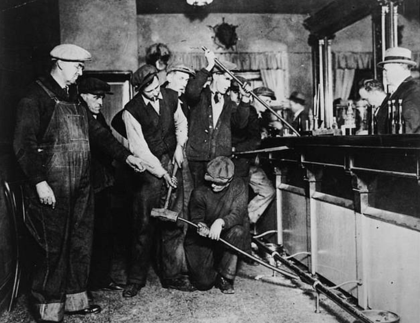 A group of men, some holding tools, gather around a long metal pipe connected to a bar. One man kneels, adjusting the pipe, while others observe. The setting appears to be a vintage bar or saloon with ornate decor.