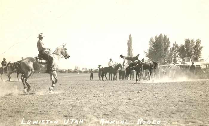 A sepia-toned photo of a rodeo in Lewiston, Utah. Cowboys on horseback are in the ring, with one horse seemingly mid-jump. Several spectators and other riders watch from the sidelines under a clear sky. Text reads "Lewiston Utah Annual Rodeo.