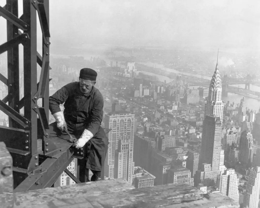 A construction worker in overalls and a cap sits on a steel beam high above a city skyline, securing bolts. The view shows many tall buildings and a distant river, with a famous skyscraper visible on the right. The scene is black and white.