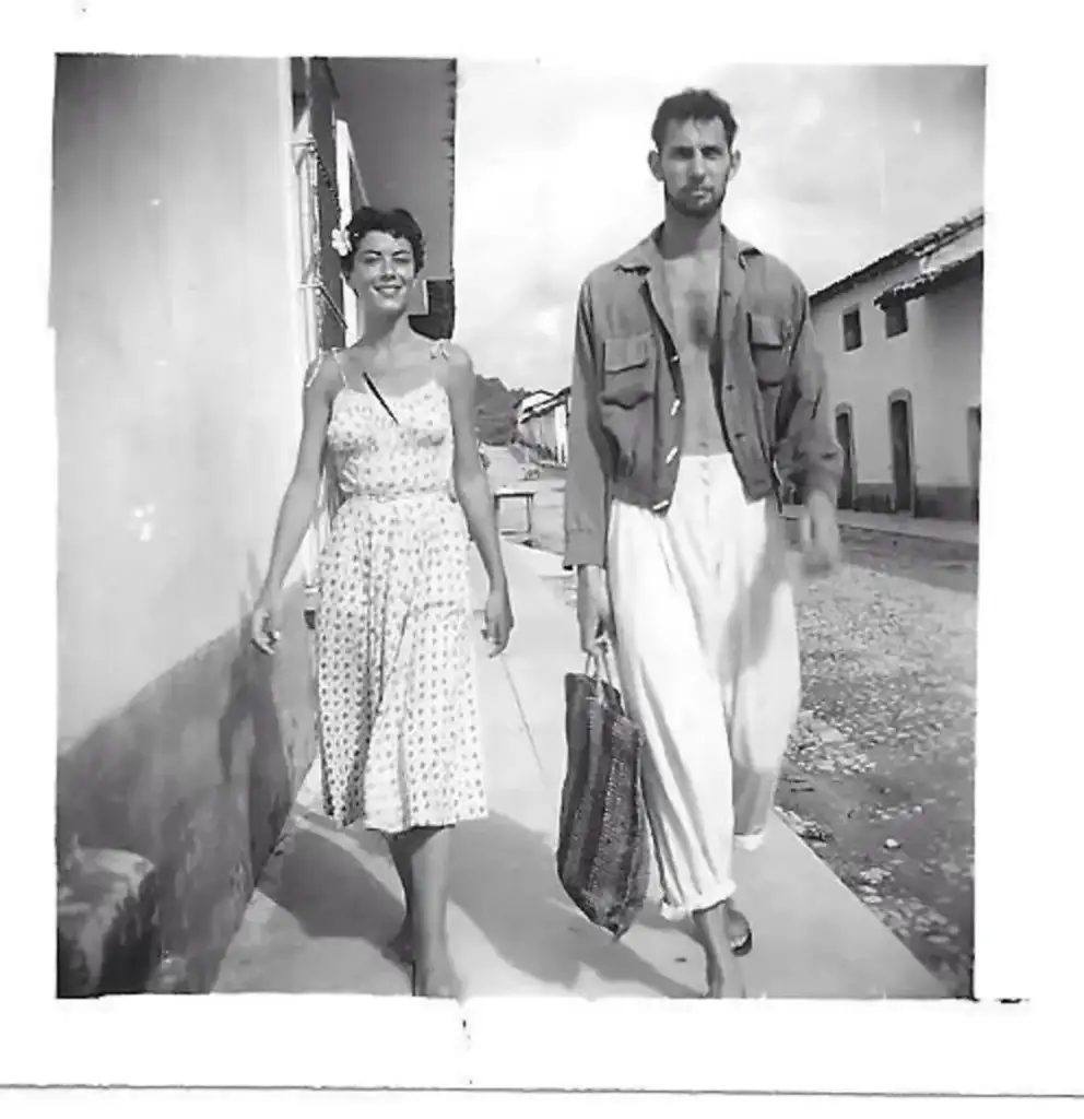 A black and white photo of a smiling woman in a patterned dress walking next to a man in a jacket with an open shirt, carrying a bag. They are on a sidewalk with buildings and a cloudy sky in the background.