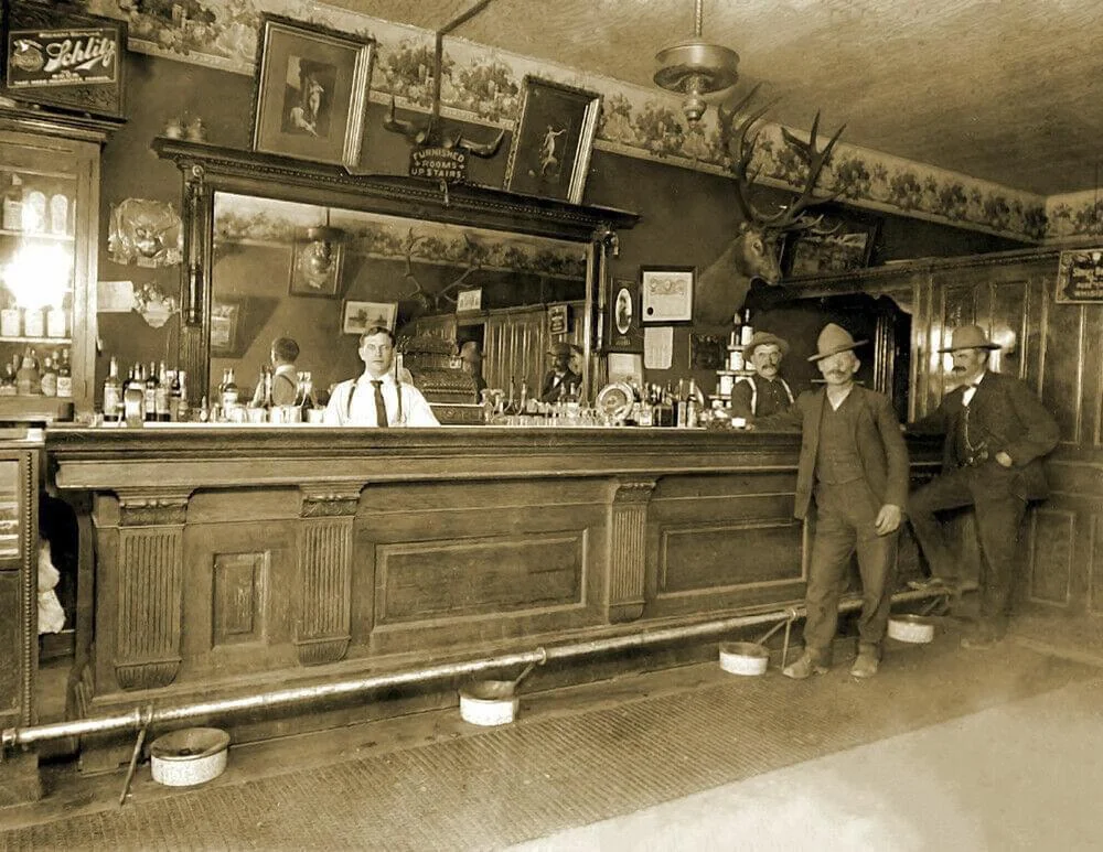 A vintage bar scene with four men in suits and hats standing around a long wooden counter. Bottles and glasses are visible on the bar. A large mirror behind them reflects the room, which is decorated with mounted animal heads and framed pictures.