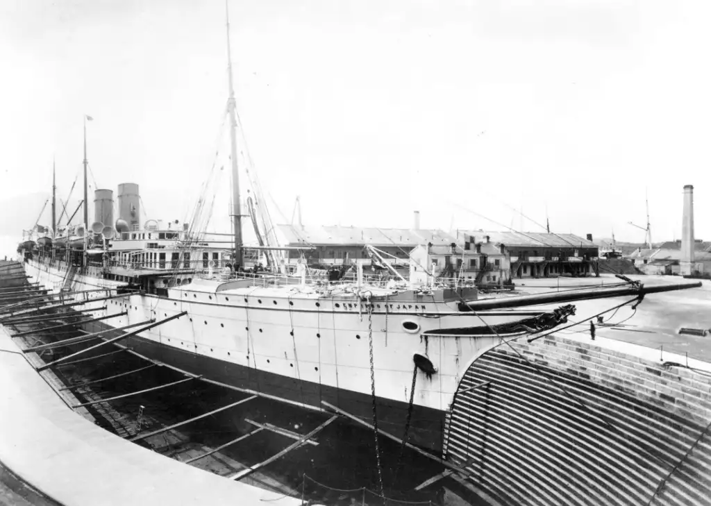 Black and white photo of a large steamship in a dry dock. The ship has multiple decks and tall masts, with visible ladders and railings. Industrial buildings are in the background, and the dock is surrounded by stone walls.