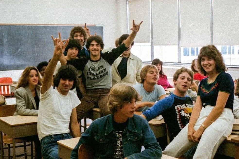 A group of smiling students casually pose in a classroom. Some are sitting on desks while others are standing. A chalkboard and window blinds are visible in the background. One person in the front holds up a hand sign.