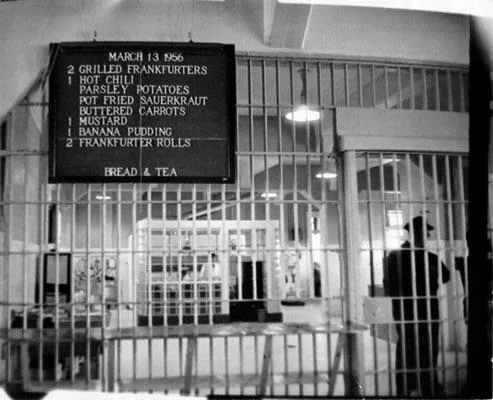 Black and white image of a menu board dated March 13, 1956, listing food items such as grilled frankfurters, hot chili, parsley potatoes, buttered carrots, mustard, banana pudding, and frankfurter rolls. The board hangs in a room with barred doors.