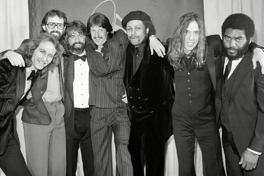 A group of seven men in formal attire, some in suits and others in tuxedos, pose together smiling for the camera at an event with a curtain backdrop. They appear to be celebrating and are in a jovial mood.