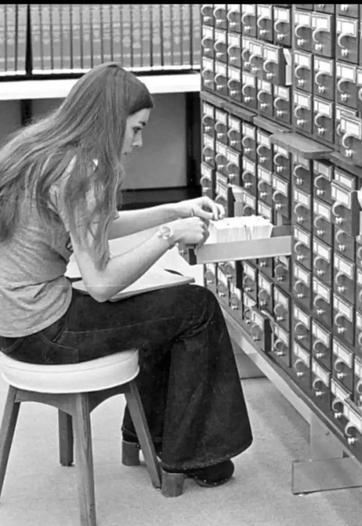 A person with long hair sits on a stool, browsing a card catalog drawer in a library. Books line a shelf above, and the setting appears to be from a past era, reflecting an older method of information organization.