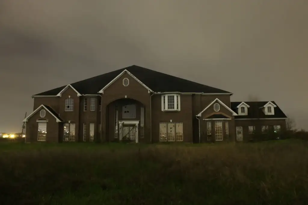 A large, dark brick mansion at night under a cloudy sky. The house has multiple windows, a prominent entrance, and a triangular roof. It sits in a grassy area, with faint lights visible in the distant background.