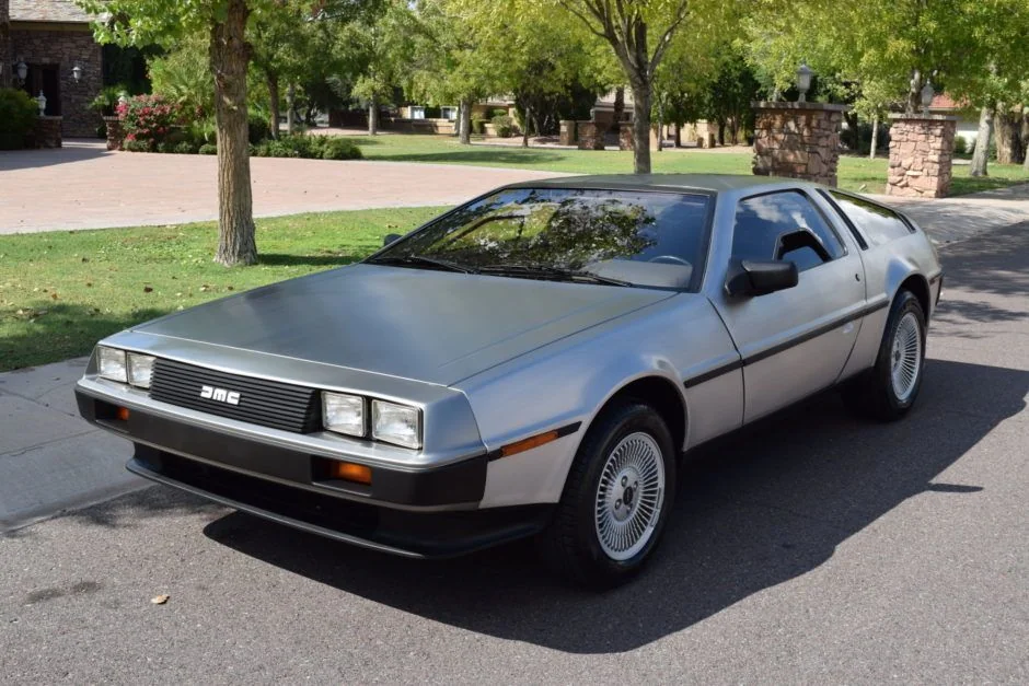 A silver DeLorean sports car is parked on a suburban street, surrounded by green trees and a residential backdrop. The car features a distinctive boxy design with gull-wing doors and a sleek, angular body.