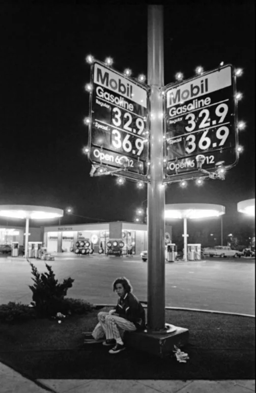 Black and white photo of a Mobil gas station at night with illuminated signs displaying gas prices of 32.9 and 36.9 cents. A person sits beneath a pole near the station. Gas pumps and a building are visible in the background.