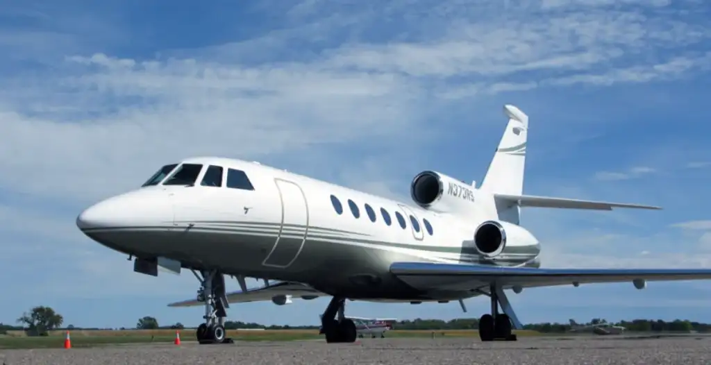 A white private jet with dark windows parked on an airport runway. The sky is blue with scattered clouds, and the runway is bordered by grass. An orange safety cone is placed nearby.