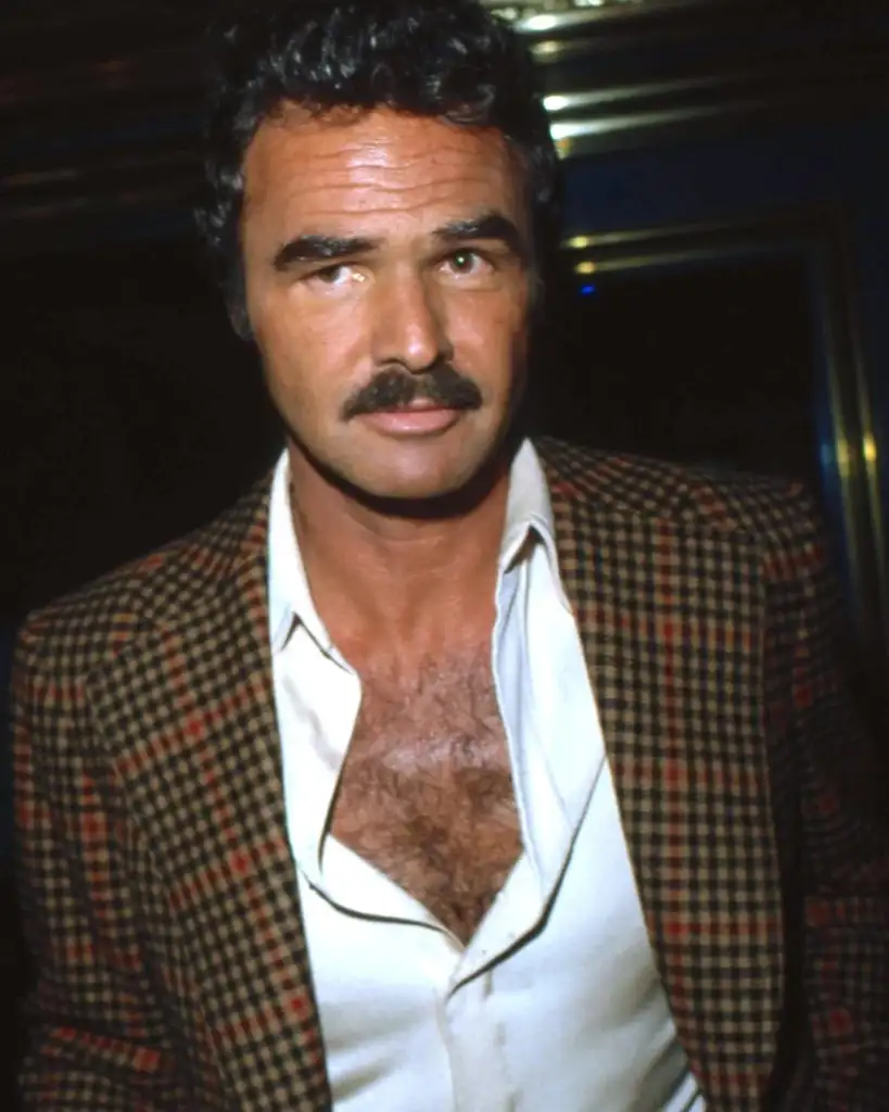 Man with a mustache, wearing a checkered blazer over an open white shirt, stands looking directly at the camera.