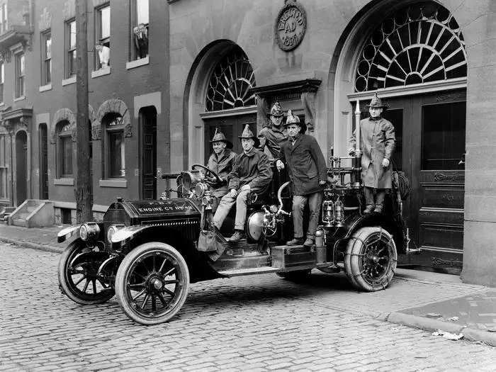 A black and white photo shows six firefighters in vintage uniforms posing with an old fire engine on a cobblestone street. The engine is parked in front of a building with large arched doorways.