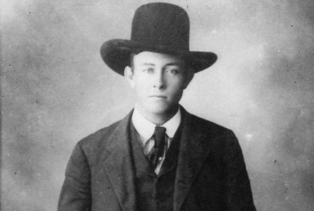 Black and white portrait of a person wearing a suit, tie, and a wide-brimmed hat. The background is plain and blurry, highlighting the figure's formal attire and serious expression.