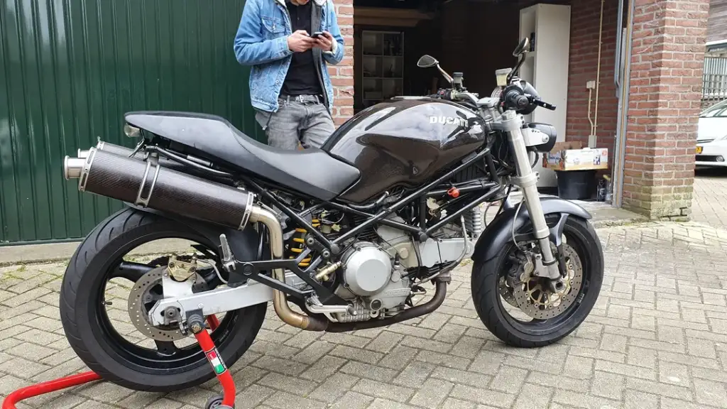 A motorcycle parked on a paved area with a person in the background using a phone. The bike has a sleek, angular design with a glossy finish and is supported by a rear stand. A brick building and garage door are visible behind.