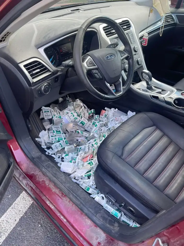 Car interior with a driver's seat, steering wheel, and dashboard of a red Ford vehicle. The floor is covered with numerous sauce packets in various colors, creating a cluttered appearance. The passenger seat is empty.