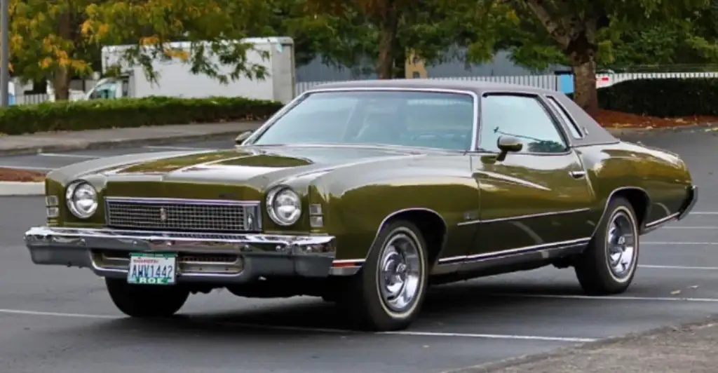 A classic green car with a brown roof is parked in an empty lot. The car has a vintage design with chrome accents and circular headlights. Trees with autumn foliage are in the background.