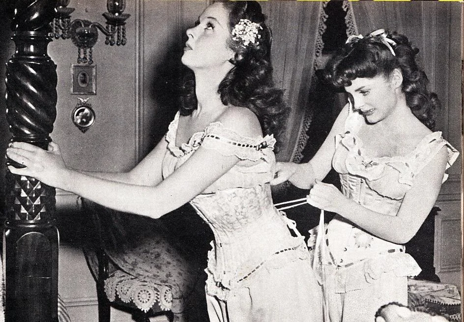 Two women in period costumes, with lace and flowers in their hair, are in a vintage setting. One is tightening a corset on the other, who leans against a bedpost. The room has ornate furniture and decorations.