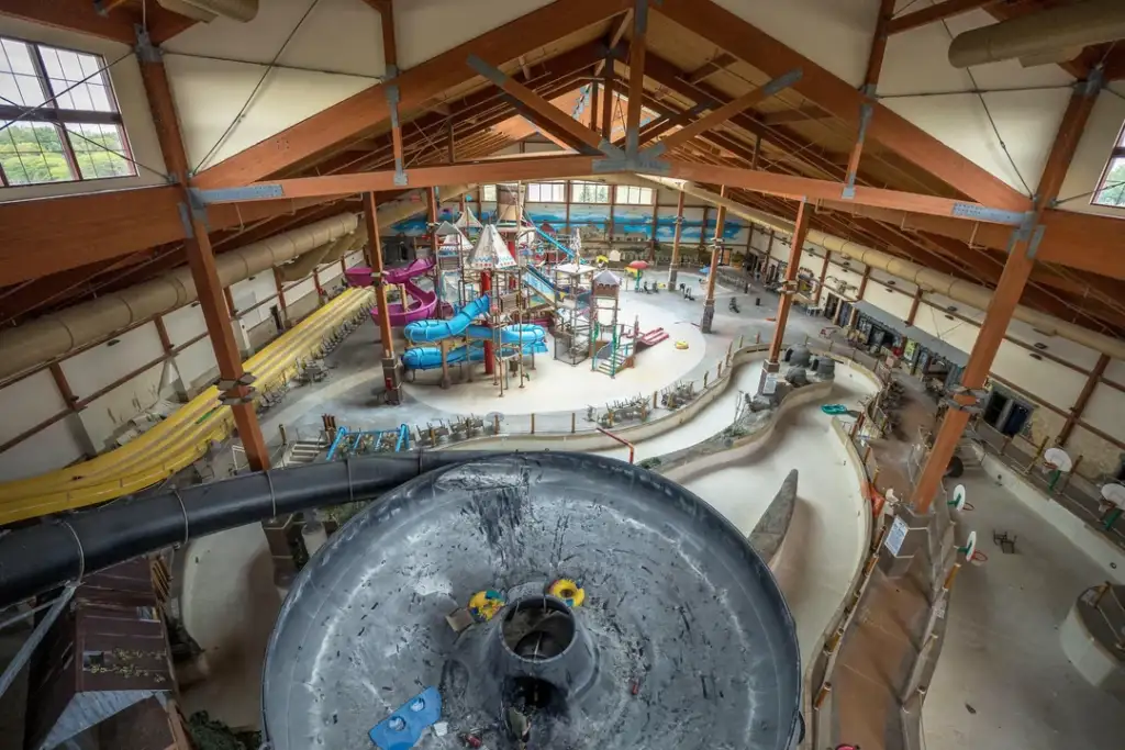 Indoor water park with multiple slides, including blue and yellow ones, and a large funnel slide in the foreground. The wooden beams of the ceiling are visible, and there are various structures and seating areas around the park.