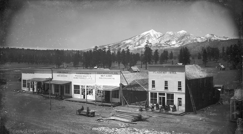 A black and white image of an old Western town with wooden buildings. Signs include "General Merchandise" and "Dresses & Suits." Snow-capped mountains and pine trees are in the background. Lumber is scattered in the foreground.