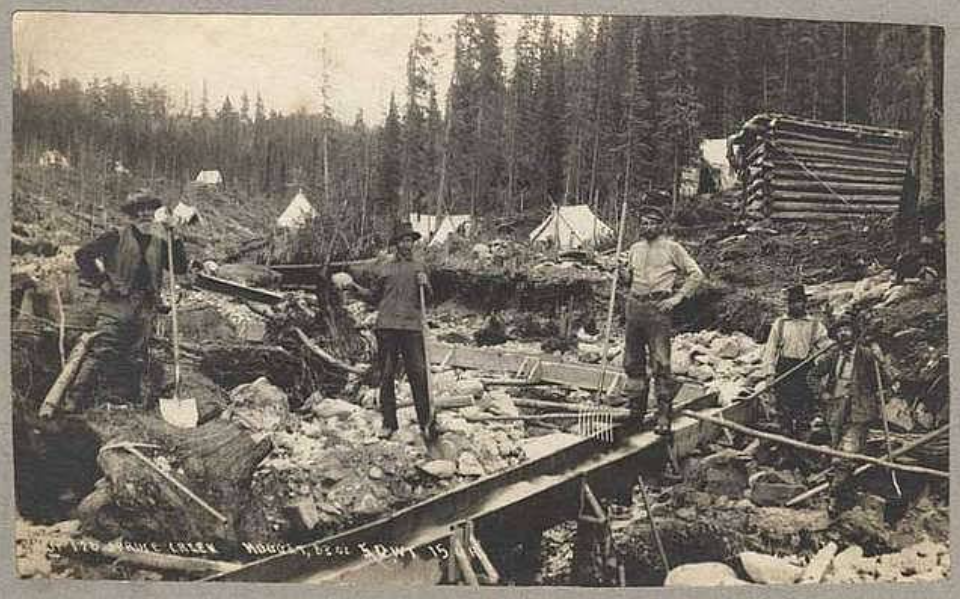 Black and white photo of five men working at a gold mining site in the woods. Some hold tools, and there are wooden structures in the background with scattered machinery and debris. Tents and trees are visible in the distance.