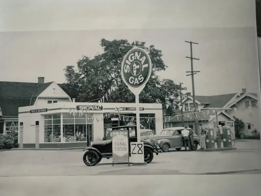 A vintage gas station with a "Signal Gas" sign stands beside a classic car parked at the pumps. The station has a small building, and trees are in the background. Triangular flags are strung overhead, adding a festive touch.