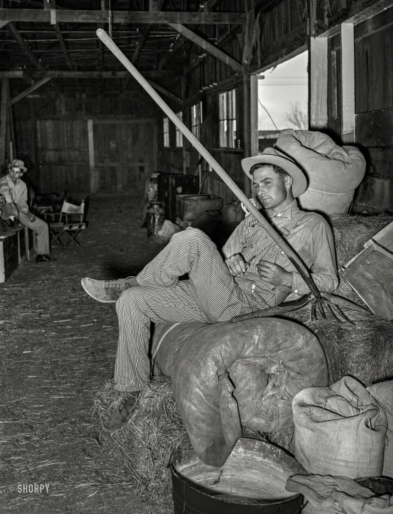 A man in striped overalls and a hat reclines on hay bales inside a barn, holding a long pitchfork. Burlap sacks are around him, and another man sits in the background on a wooden chair, watching. The scene is dimly lit with open barn doors.