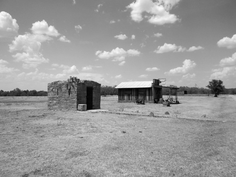 A black and white image of a rural landscape with two rustic buildings. A stone structure stands on the left, and a wooden cabin with a metal roof is on the right. The sky is partly cloudy, and the ground is grassy and expansive.