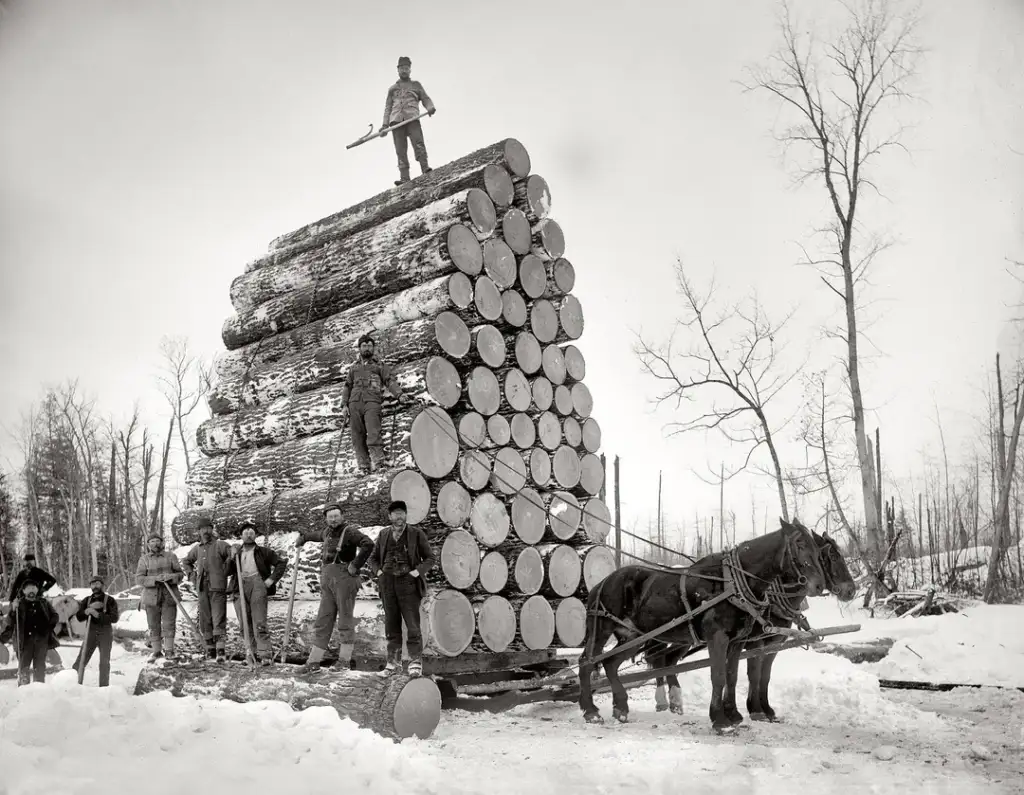 A black and white photo shows a large stack of logs on a sled pulled by two horses. Seven men stand on and around the logs, with one man on top holding an ax. Snow covers the ground, and leafless trees are in the background.