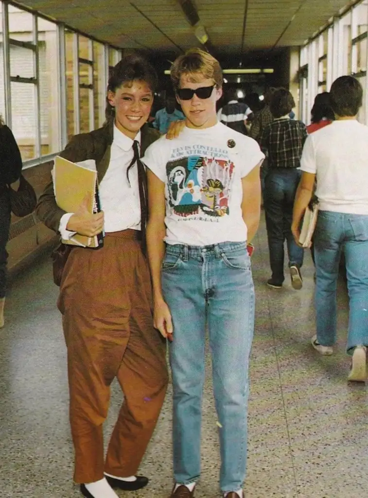 Two people stand in a school hallway. One wears a white blouse and brown pants, holding a folder. The other wears sunglasses, a graphic t-shirt, and jeans. Other students walk in the background.