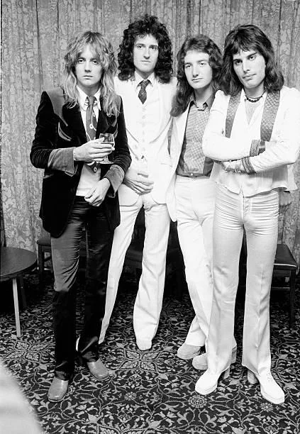 Four men in 1970s-style clothing pose together in a room with patterned carpeting and curtains. One holds a drink, and they all have long hairstyles. Their outfits include suits and flared pants, with a mix of light and dark colors.