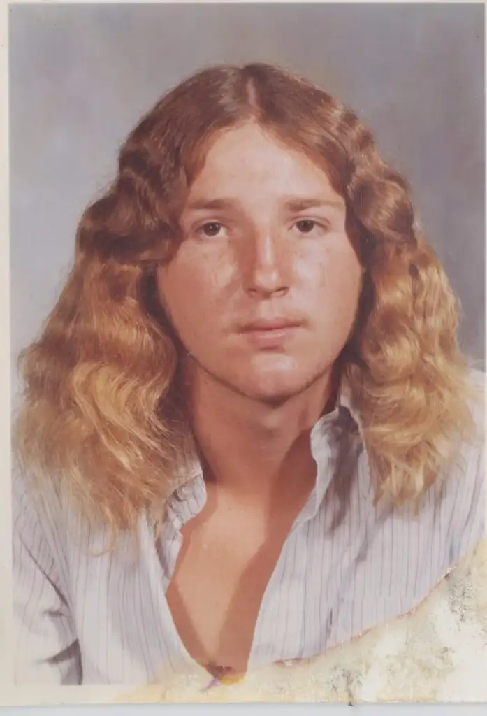 A young person with long, curly hair poses for a portrait in a light-colored, striped shirt. The photo has a slightly vintage appearance with some visible wear at the edges.